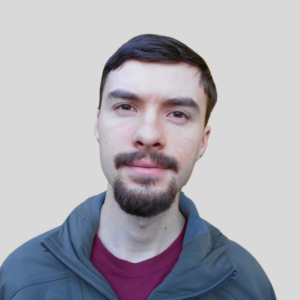 Молодой человек с короткими темными волосами и козлиной бородкой, одетый в повседневный пиджак поверх бордовой рубашки.<br />
У него нейтральное выражение лица, и он смотрит прямо в камеру. 