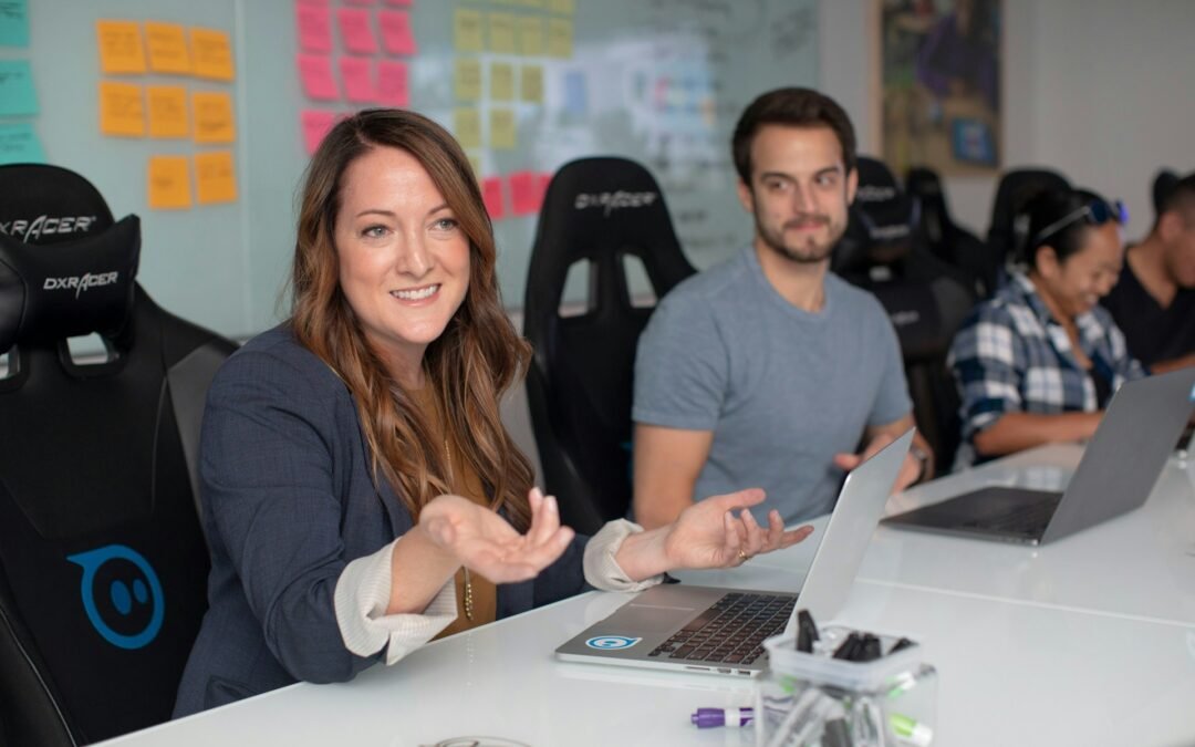 Team members collaborating in a modern office space, with a woman leading the discussion at a desk with laptops and colorful sticky notes in the background.