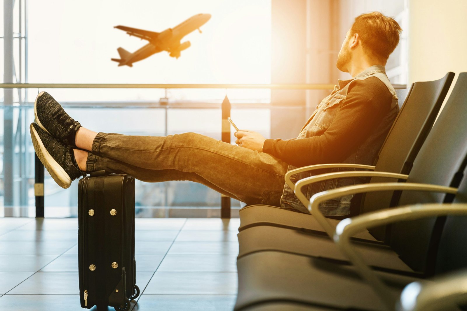 Traveler at an airport lounge with feet resting on a suitcase, looking at an airplane taking off in the background.