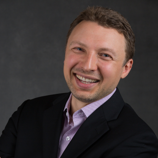 Professional headshot of Sergey, Chief Operating Officer at Stelmakh & Associates, wearing a black blazer and light purple shirt, smiling confidently against a dark gray background.