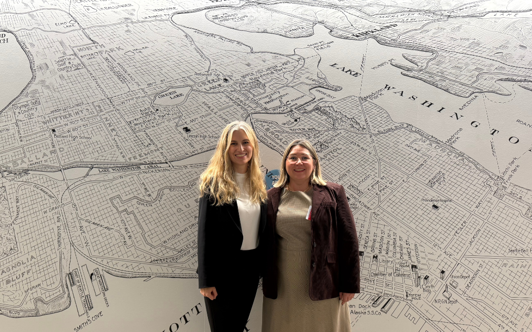 Katya Stelmakh and Olga Prygoda standing in front of a large black and white map of Seattle, smiling and dressed in business attire.